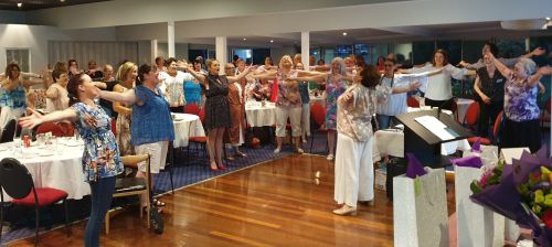 A group of mature women wearing activewear, smiling with arms wide.