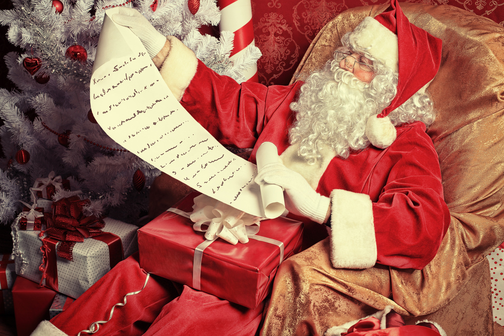 Santa Claus looks at a very long wish list. He is surrounded by presents and a Christmas tree.