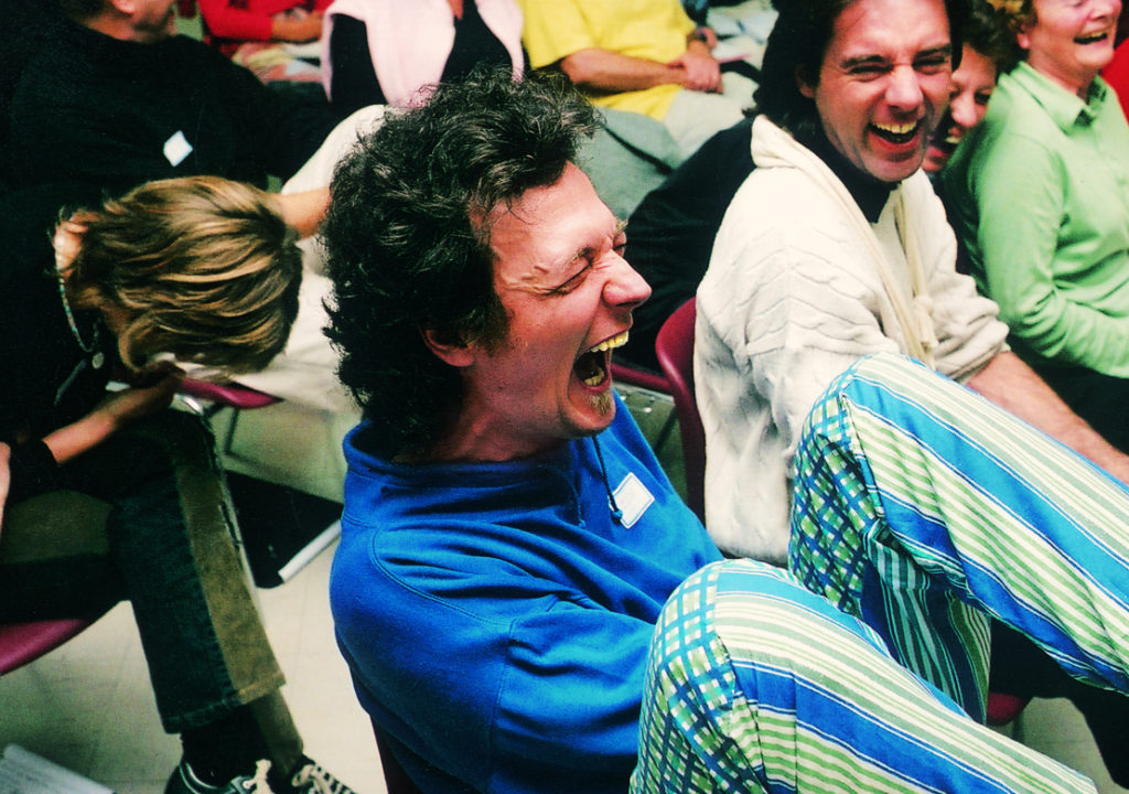 A man sitting down surrounded by people in a room all cracking up laughing