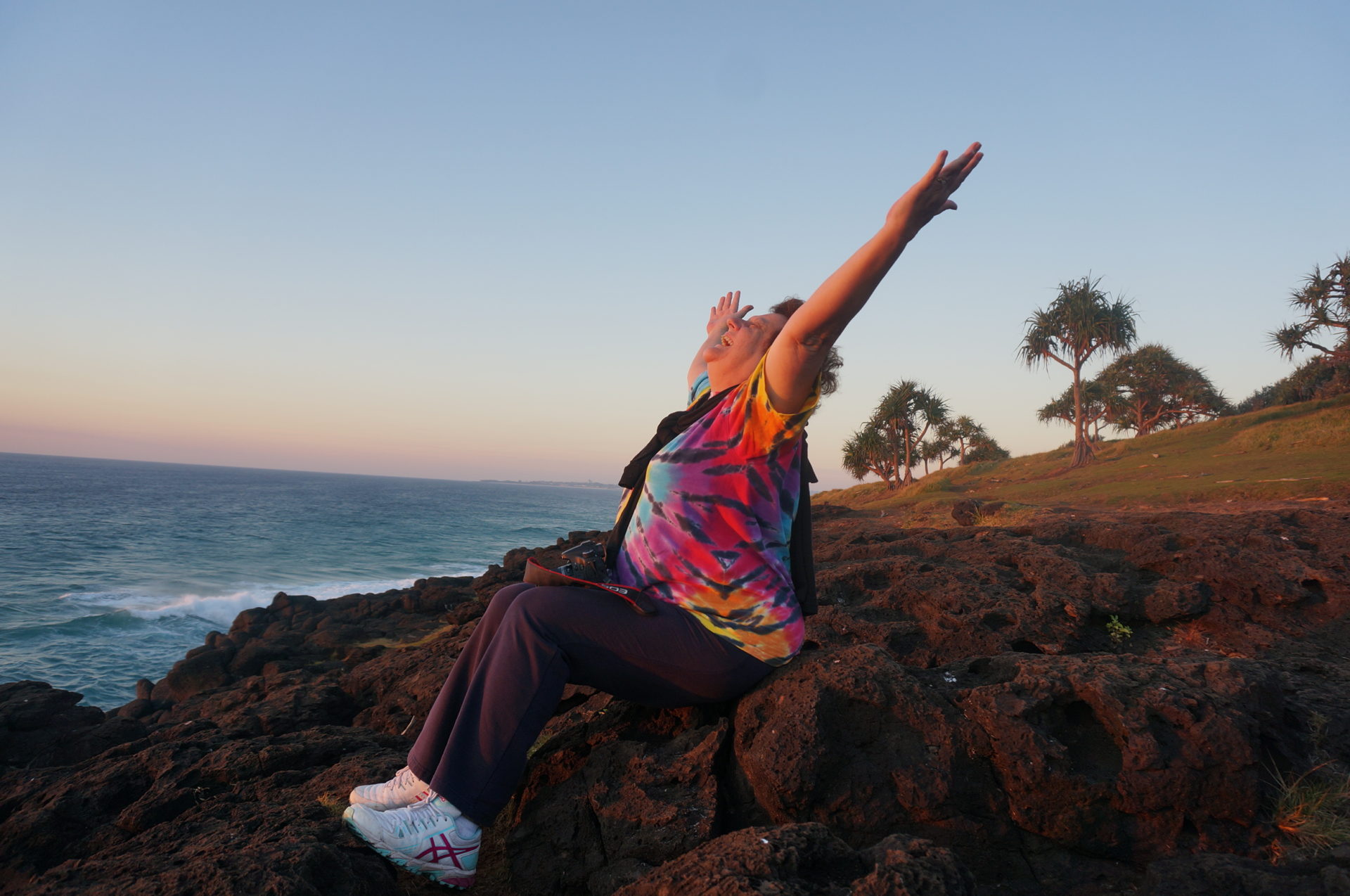 Laughter yoga facilitator Heather Joy Campbell stretches and laughs sitting on rocks in front of the ocean in sunset