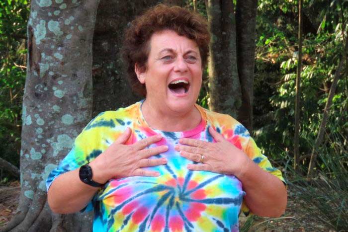 Heather Joy wearing bright rainbow top with hands on chest standing outdoors in nature.