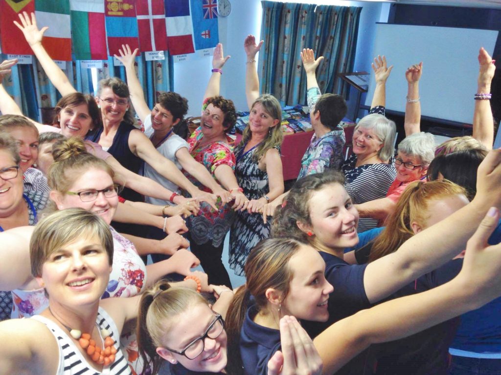 A group of women of all ages standing together one behind the other in a circle with inner hands connected by thumbs and outer hand held up