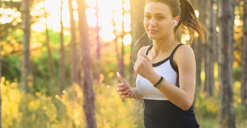 Young lady running with headphones in front of sunlit trees