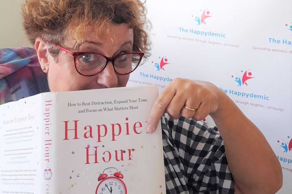 A woman holds a book called Happier Hour by Cassie Holmes. She looks knowingly at the book.