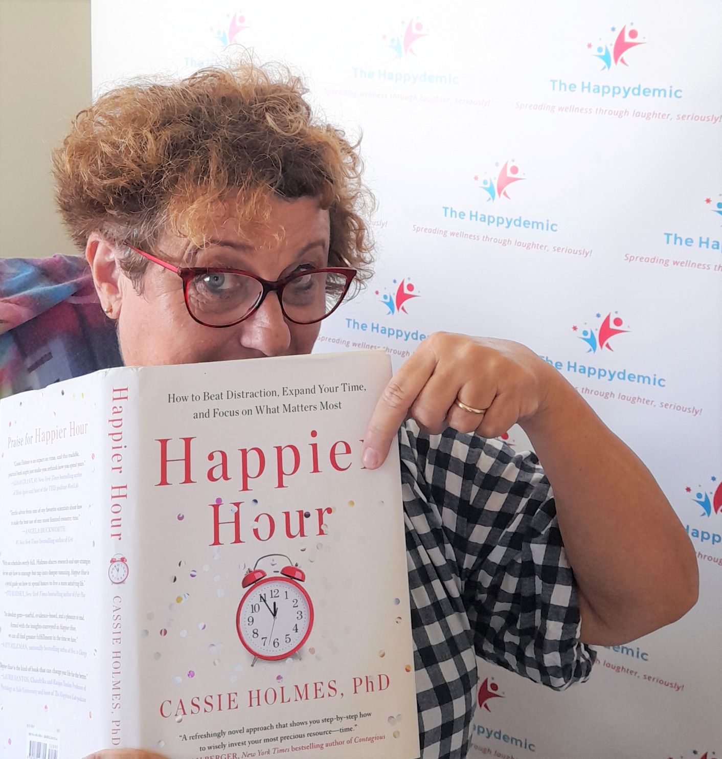 A woman holds a book called Happier Hour by Cassie Holmes. She looks knowingly at the book.