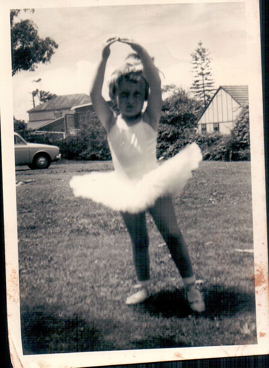 A little girl wearing a ballet tutu with arms extended over her head and one leg extended to the front, pointing her toes. She is learning ballet.