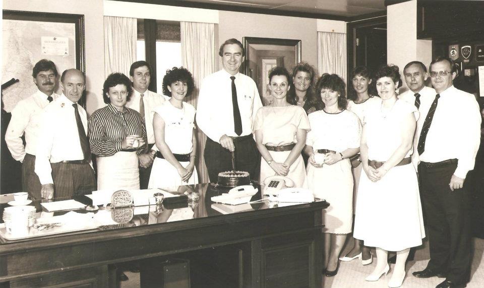 HeatherJoy is third from the left in a group staff photo. She looks serious and tired. She was working as a media advisor and speech writer for Queensland Premier Mike Ahern. he is cutting a cake on his first anniversary in the job.