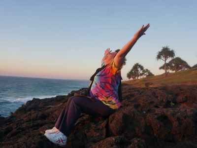 Laughter yoga facilitator Heather Joy Campbell stretches and laughs sitting on rocks in front of the ocean in sunset