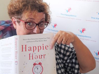 A woman holds a book called Happier Hour by Cassie Holmes. She looks knowingly at the book.
