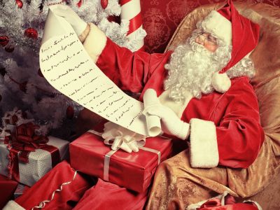 Santa Claus looks at a very long wish list. He is surrounded by presents and a Christmas tree.